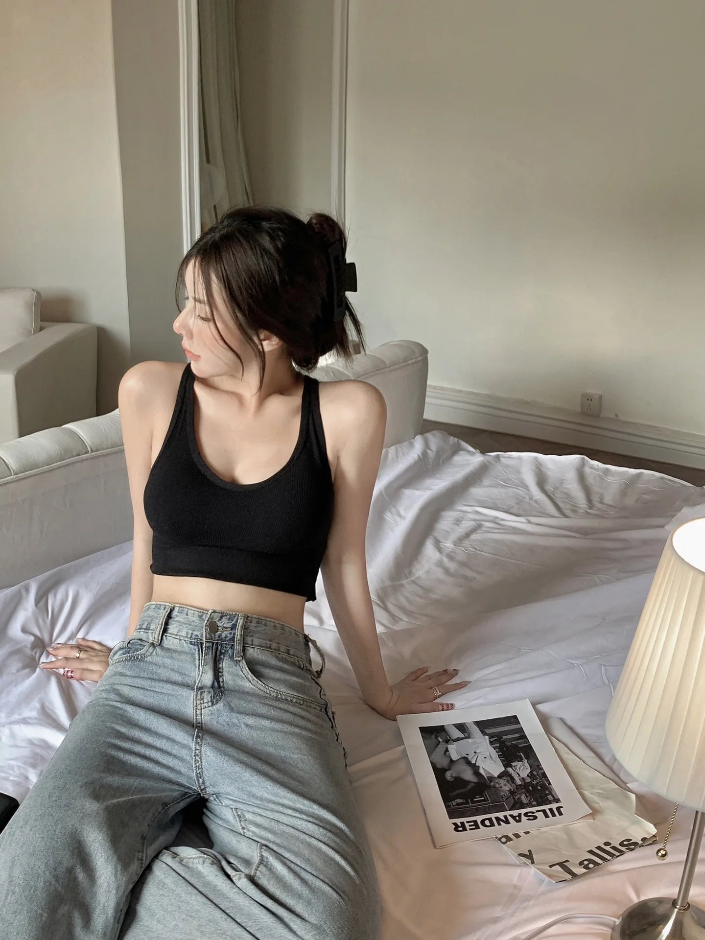 Woman sitting on bed wearing black push up sports bra sportswear crop top and high waisted jeans in cozy bedroom
