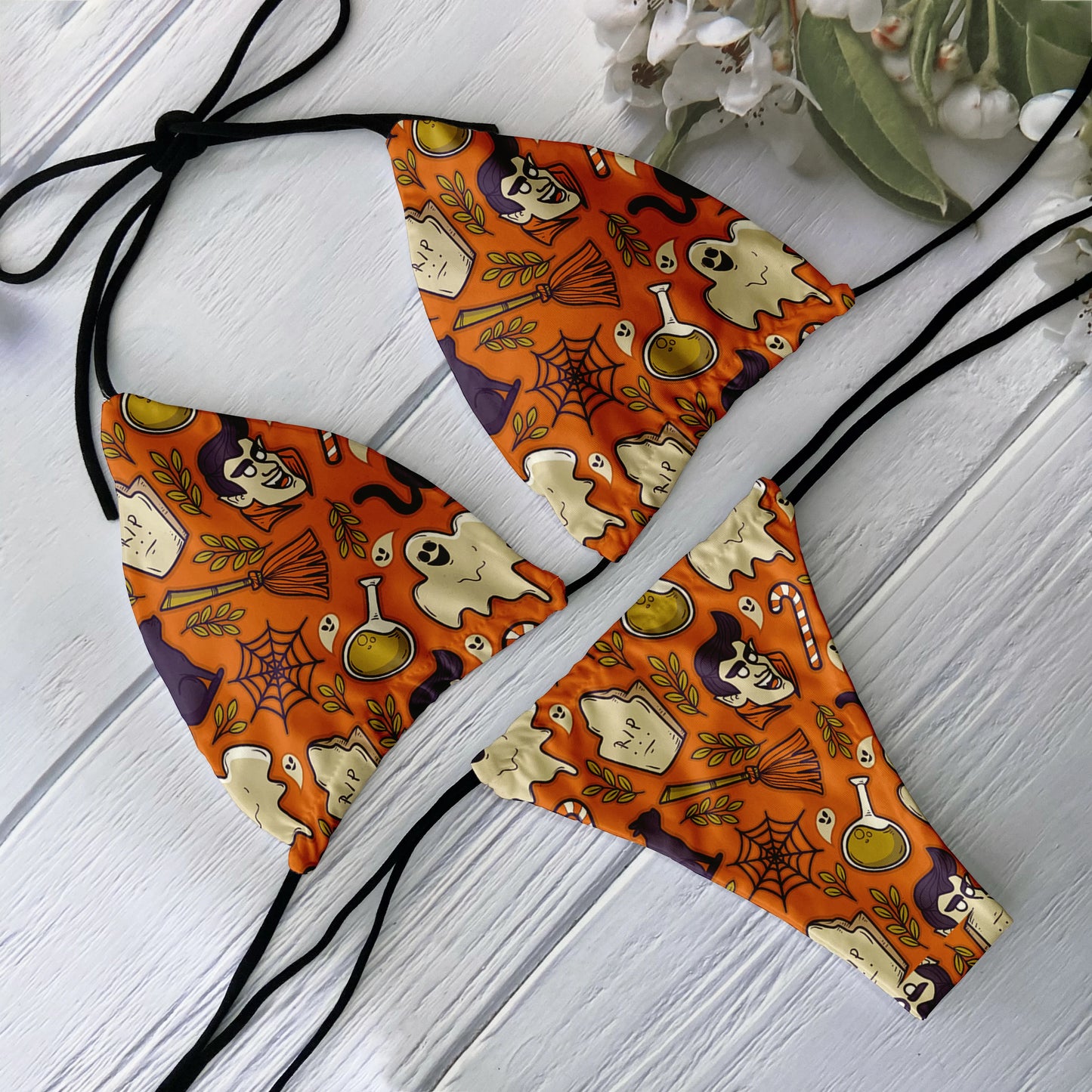 Orange Halloween themed skull print bikini set with ghosts potions cobwebs witch hats and black tie strings on white wood board
