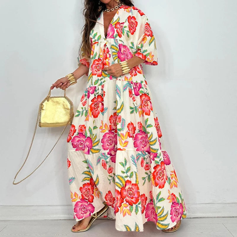 Model wearing elegant printed v-neck maxi dress in cream with bold pink floral print, tiered skirt, short sleeves and gold clutch