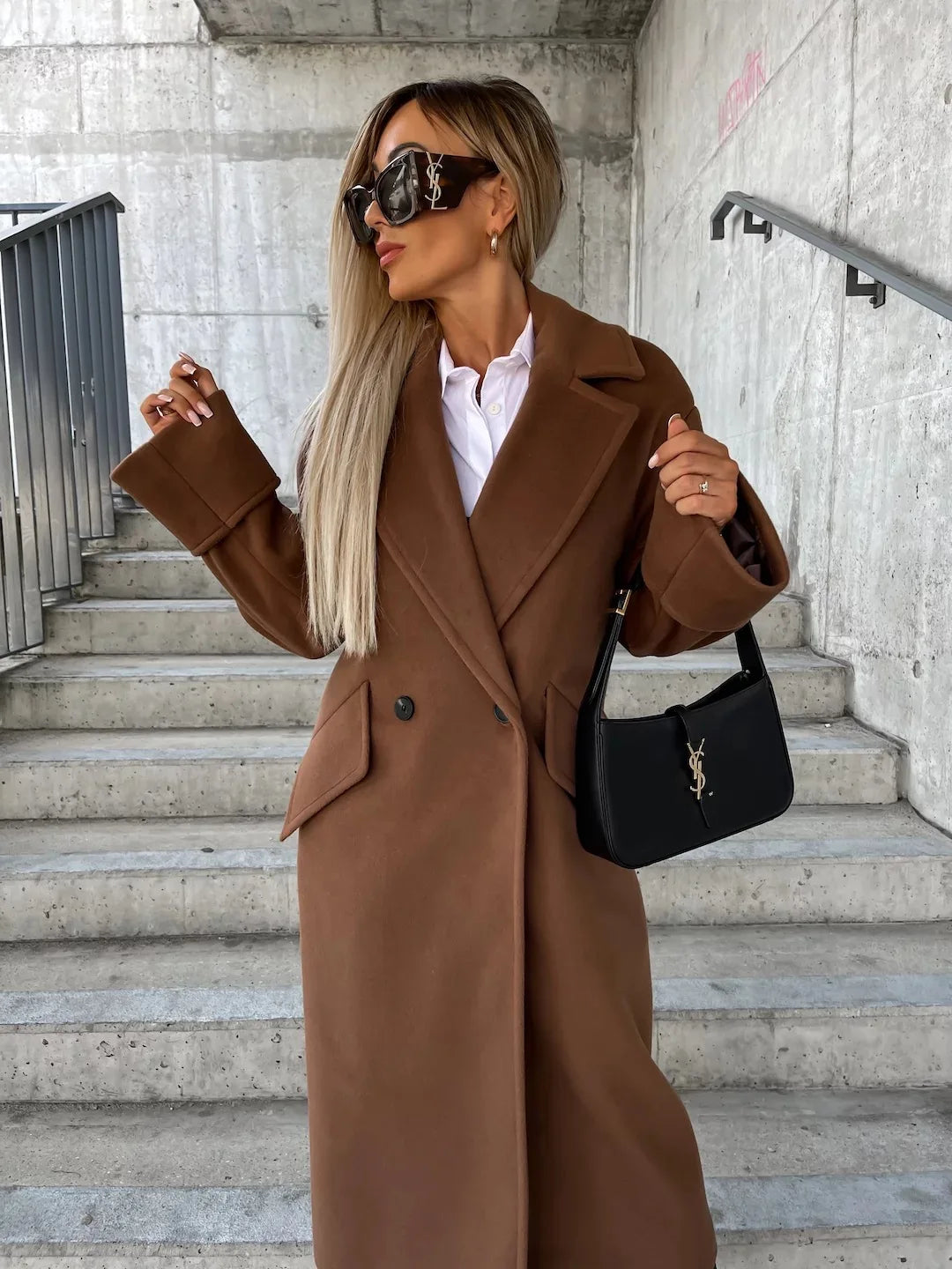 Model wearing a brown double breasted elegant long coat for women with white shirt black shoulder bag on concrete stairs