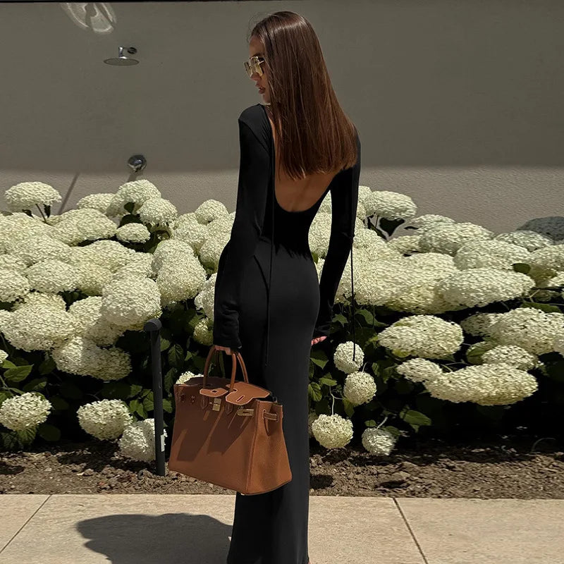Model wearing an elegant off shoulder evening dress in black, showcasing an open back and a stylish handbag