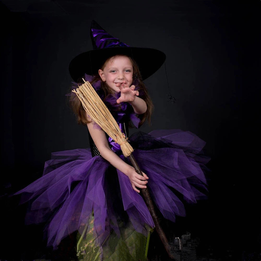 Cute girl in a kids witch tutu costume with hat, holding a broom and striking a playful pose