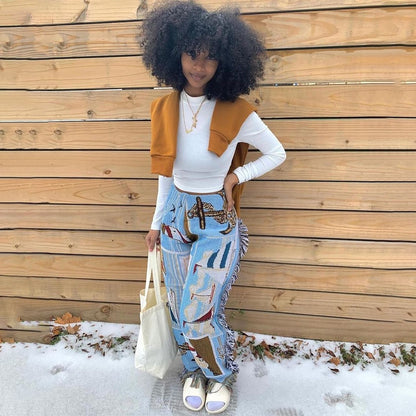 Model wearing tassel patchwork striped pants with blue panels and side tassels white crop top and mustard sweater